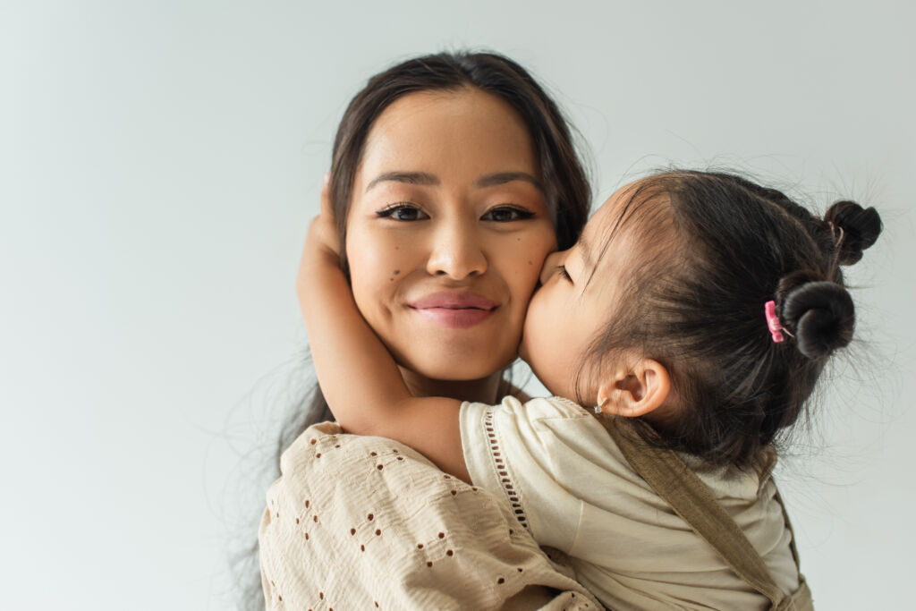asian toddler girl kissing cheek of happy mother isolated on gre