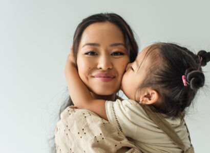 asian toddler girl kissing cheek of happy mother isolated on gre